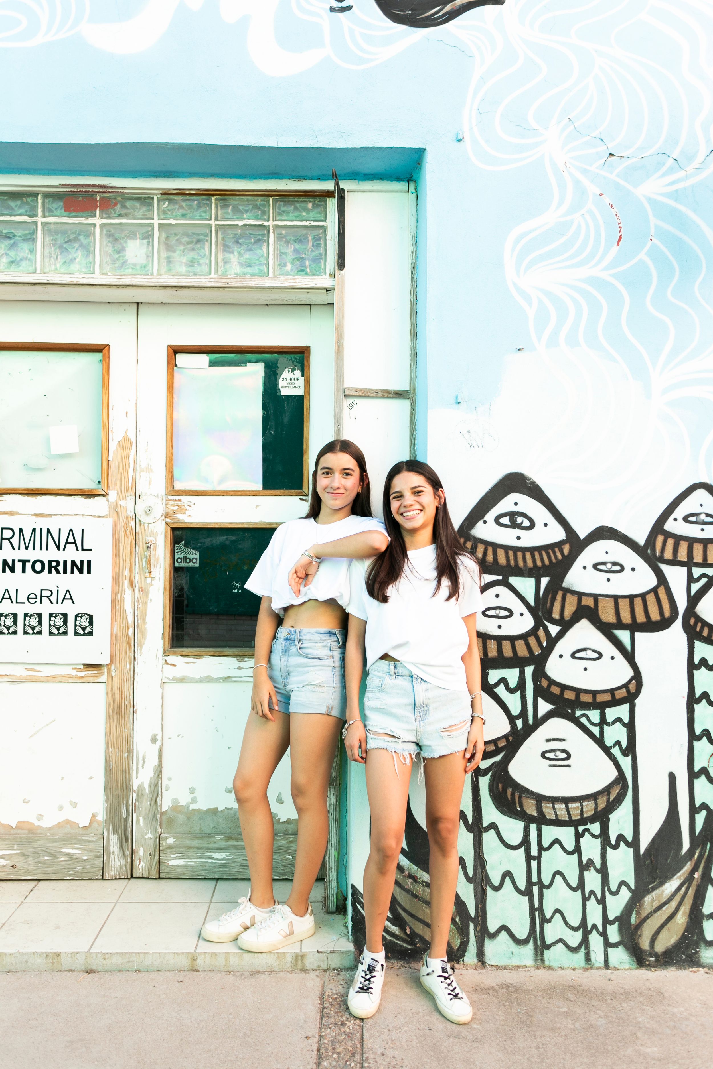 Two friends posing in casual summer outfits against wall with mushroom street art.
