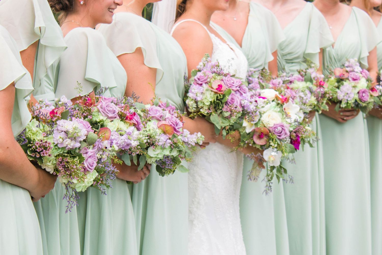 Bridesmaids in mint green dresses hold lush pink and purple floral bouquets at a springtime wedding ceremony.