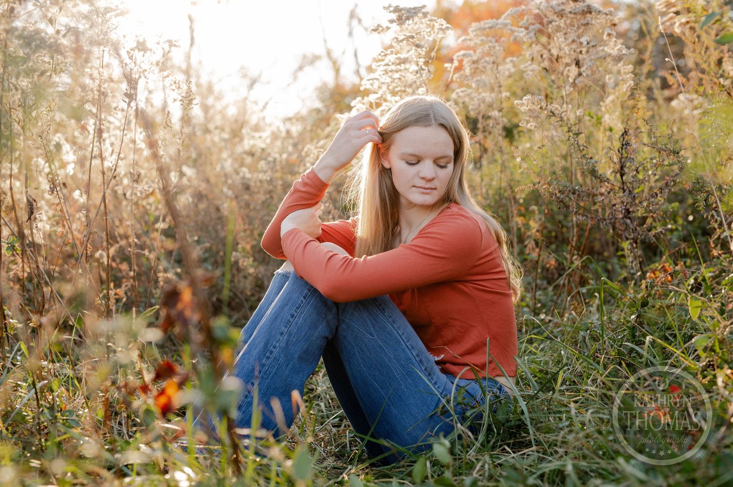 Natural light portrait in orange sweater sitting among wild autumn grasses at sunset.