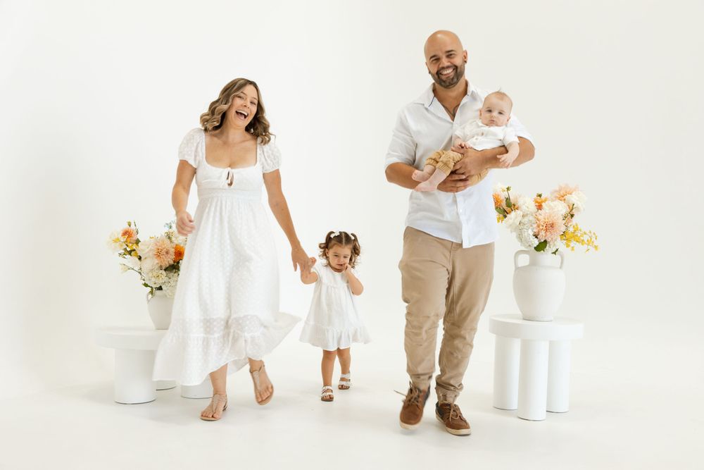 Family in coordinated white outfits poses together against bright studio backdrop with floral accents.