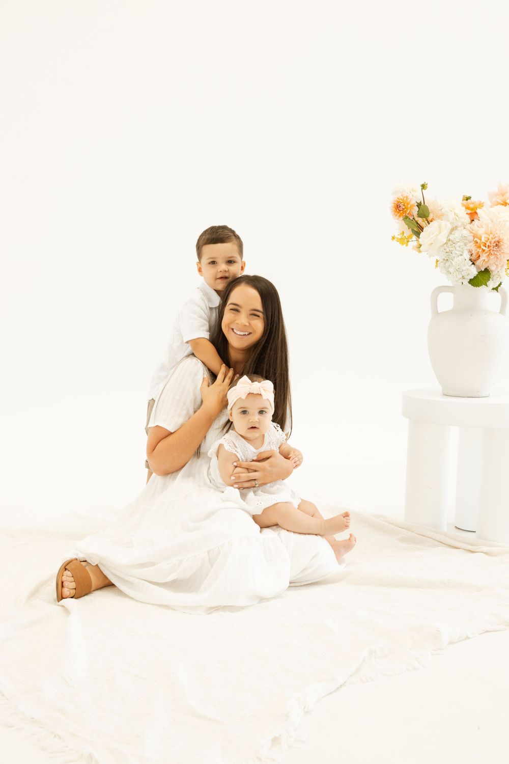 Mother in white dress holds two children against minimalist white backdrop with flowers.