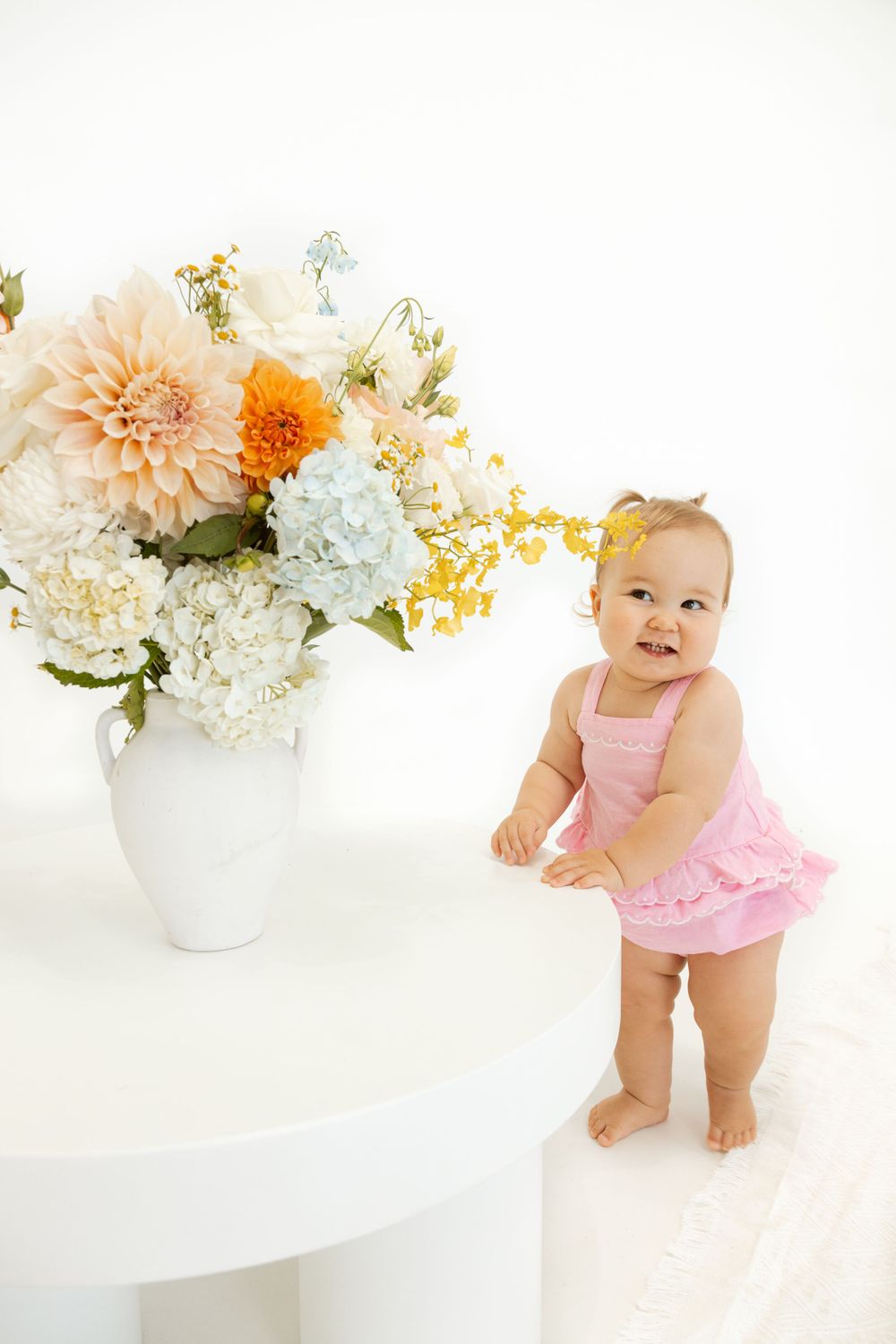 Baby in pink outfit stands next to elegant floral arrangement on white table.