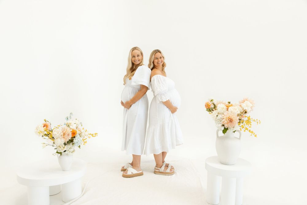 Two people in white dresses pose together against bright backdrop with floral arrangements.