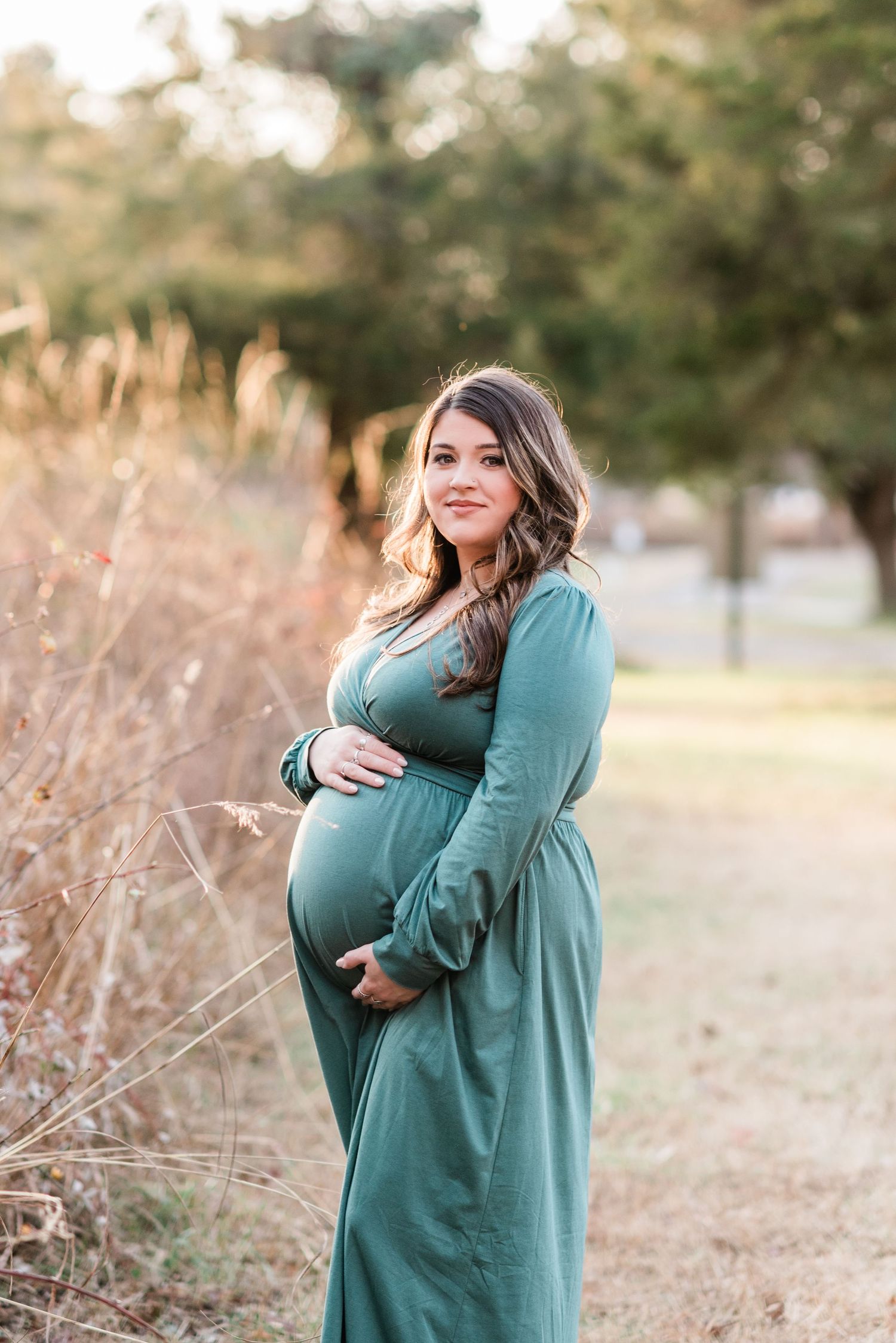 Pregnant woman in teal maxi dress poses in golden field during sunset maternity photoshoot.