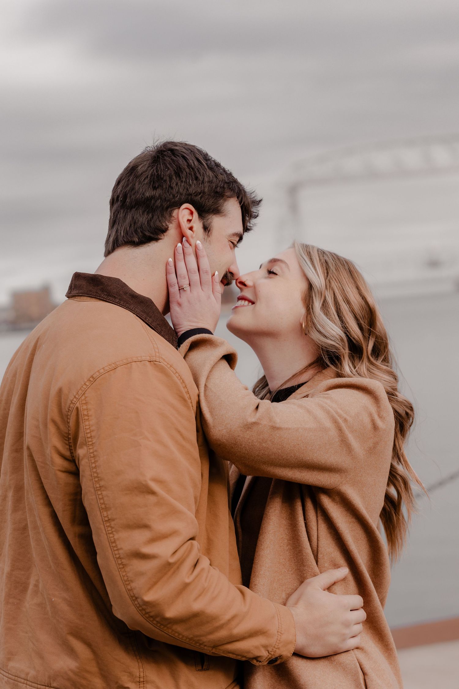A romantic couple in brown jackets share an intimate embrace and kiss against a cloudy sky background. Duluth, MN