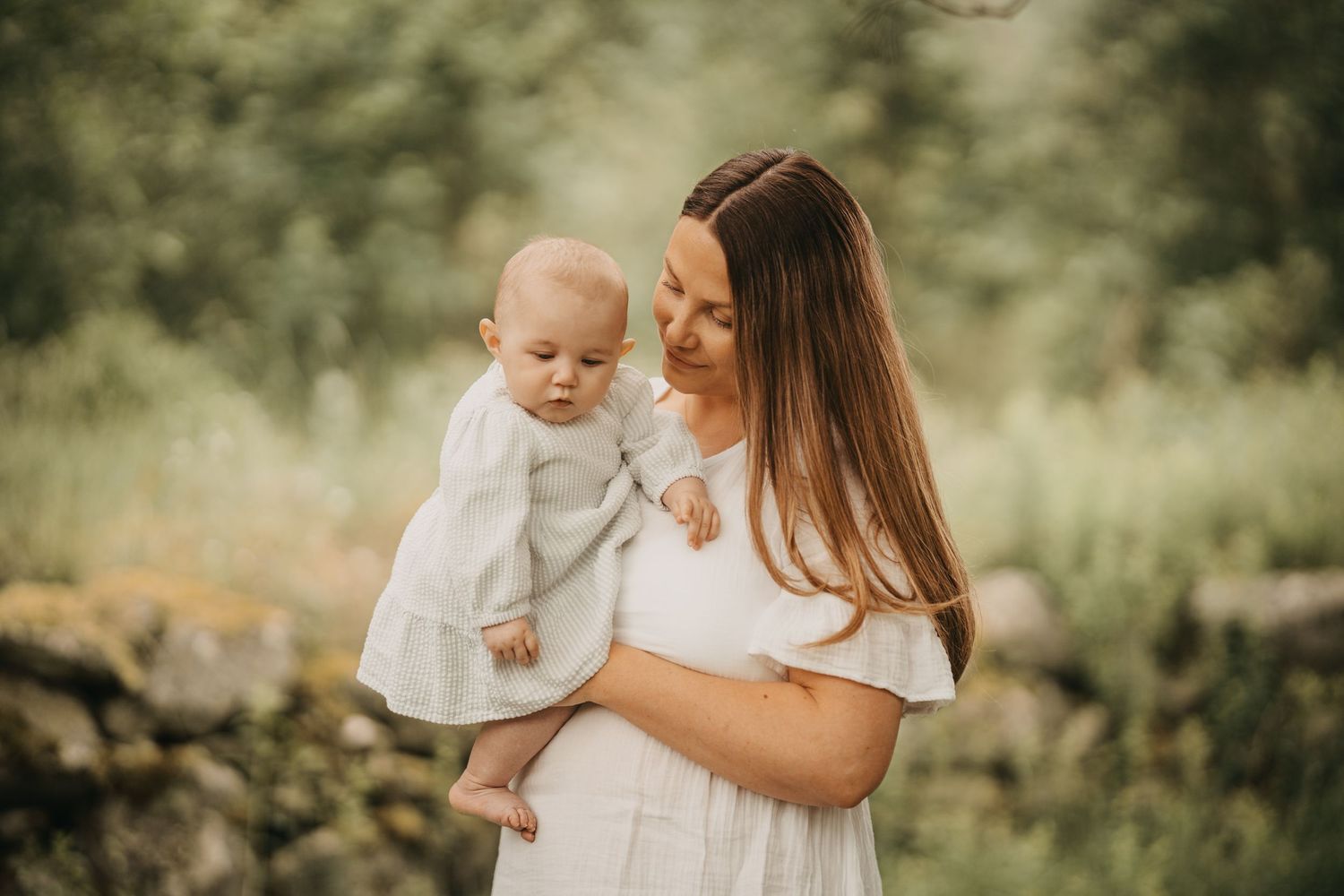 A tender moment captured in warm sunlight as parent and baby in white outfits share an intimate outdoor embrace.