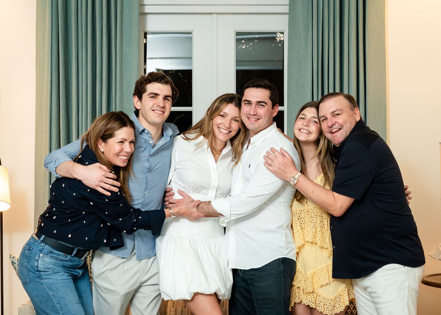 Happy family of six smiling and embracing together indoors in a warmly lit room.