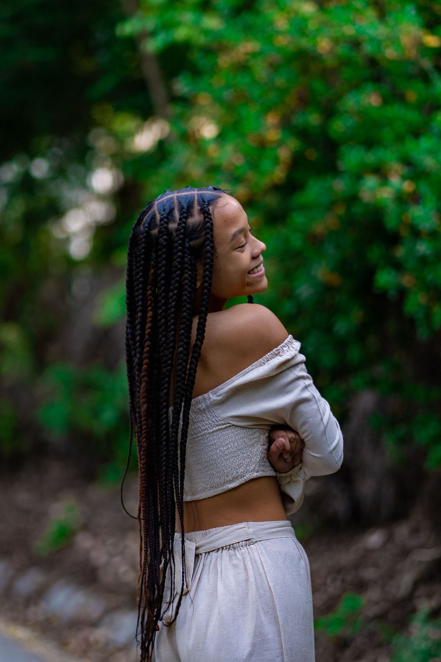 A person with long box braids poses in a white crop top and skirt against a blurred green natural backdrop.
