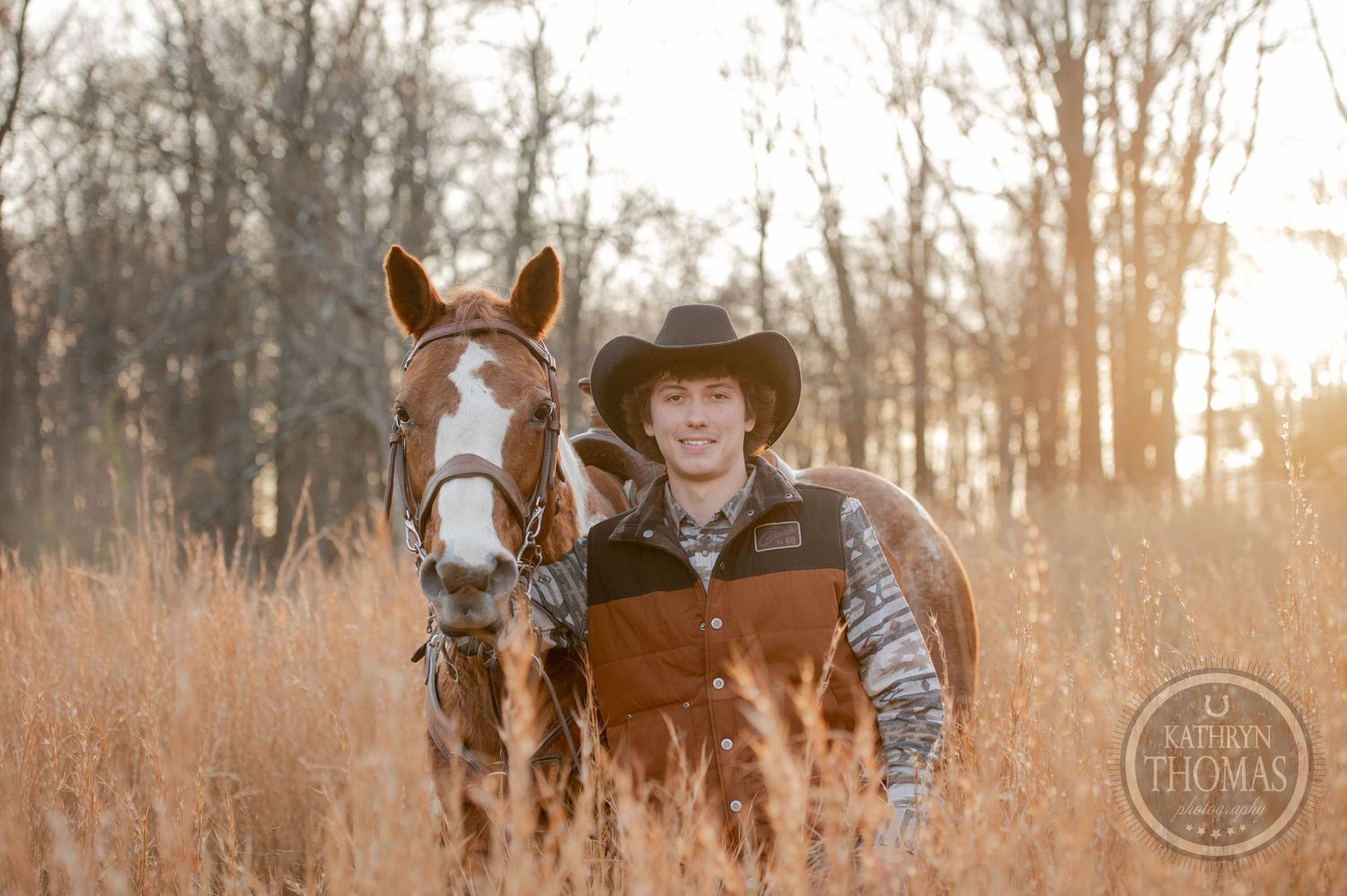 A rider in western wear stands with their paint horse in tall autumn grass at sunset.
