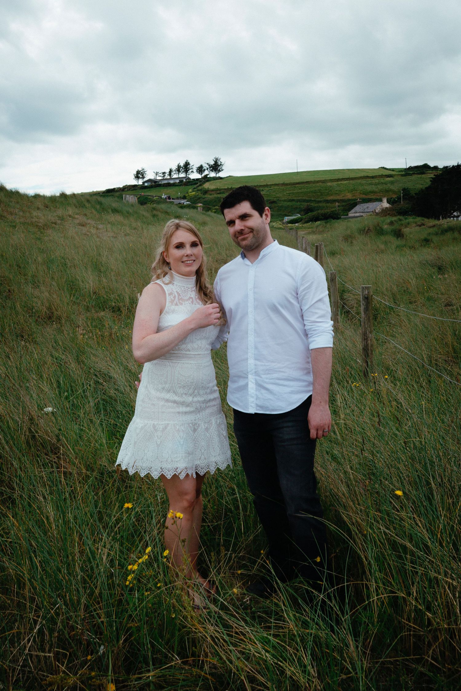 A couple poses together in a grassy meadow with rolling hills and moody clouds in the background.