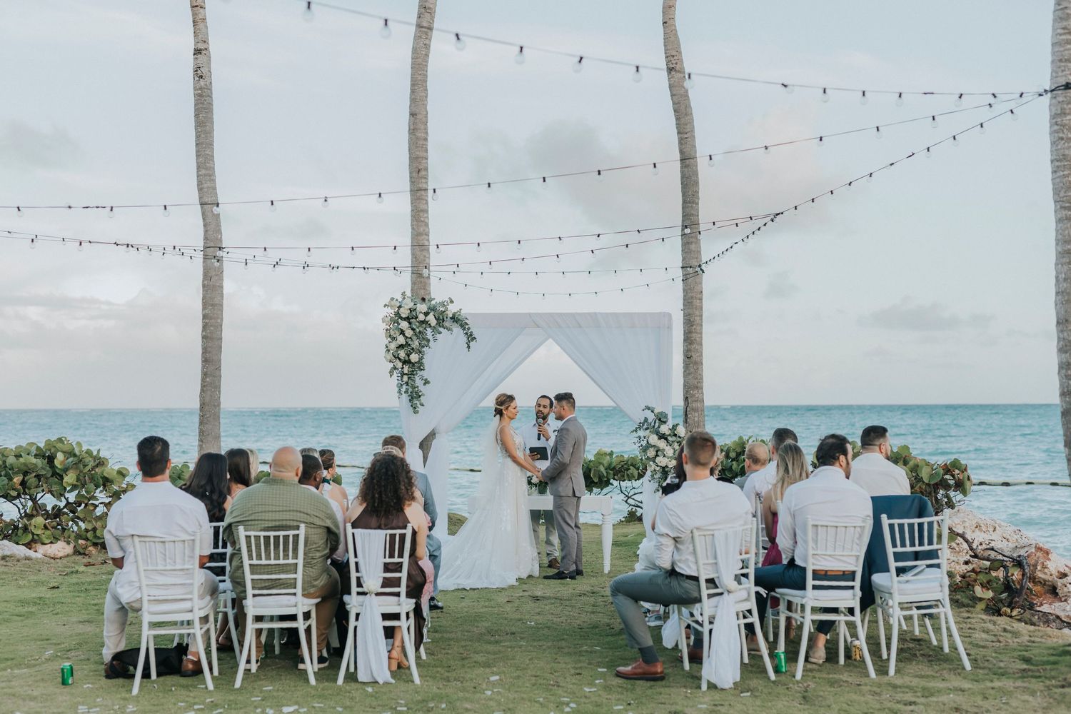 An intimate beachside wedding ceremony with white chairs, string lights, and turquoise ocean views in the background.