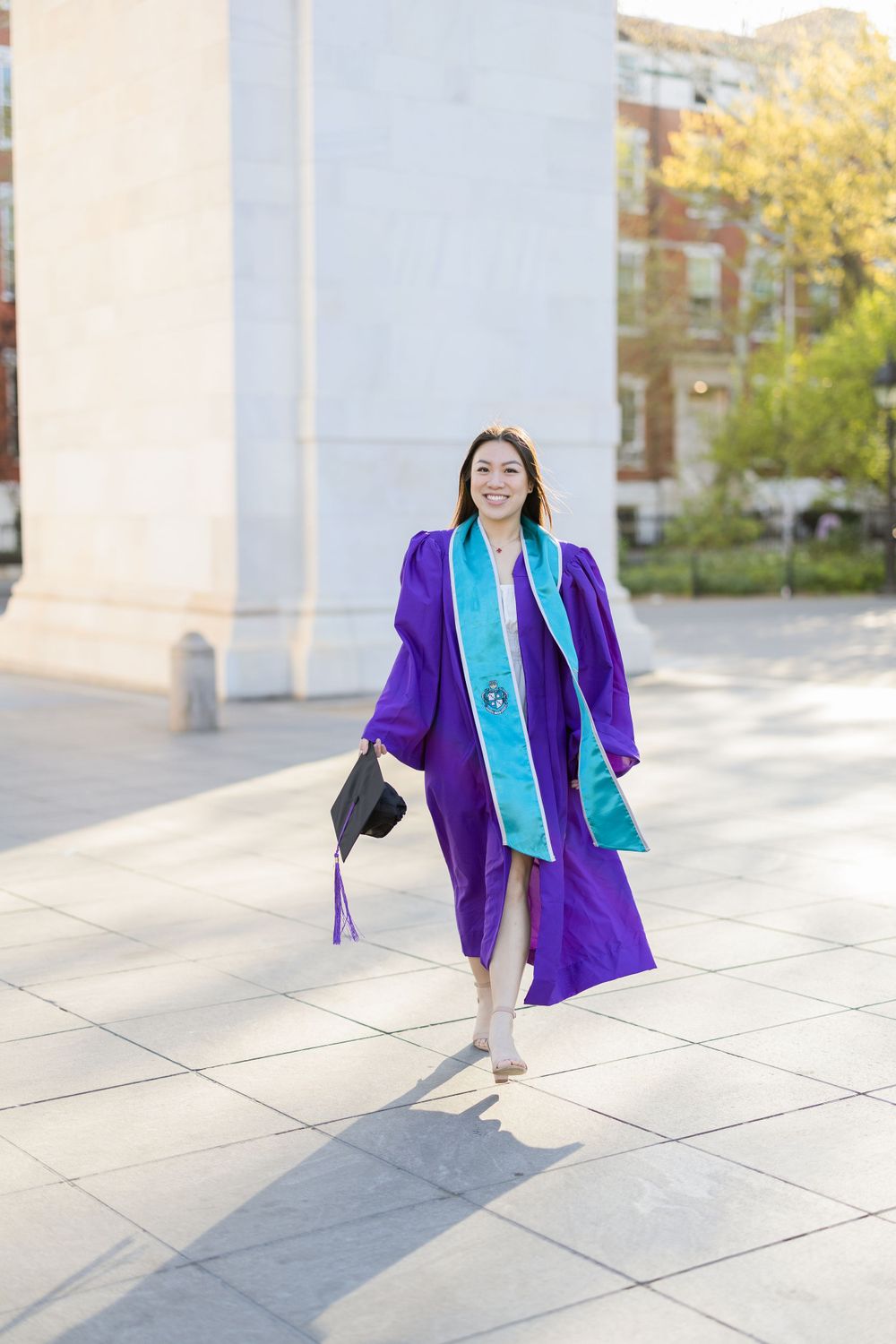Graduate in purple graduation gown and turquoise stole walking with diploma near stone wall.