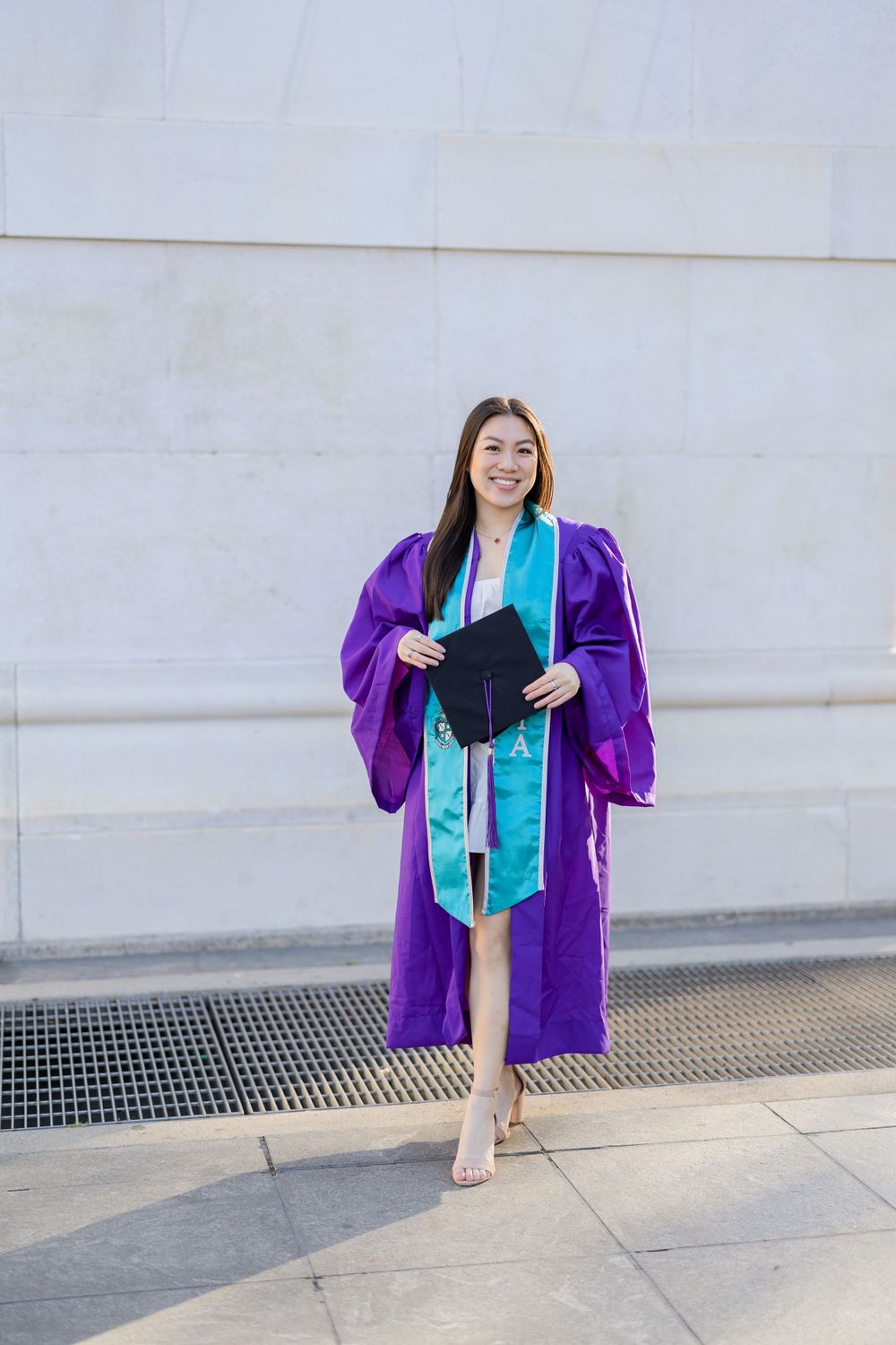 Graduate holding diploma while posing in purple gown with turquoise stole against white wall.