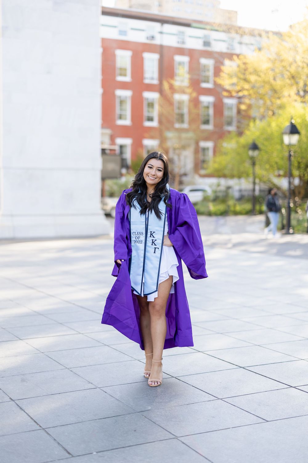 Graduate in purple graduation gown and light blue stole walking on campus sidewalk.