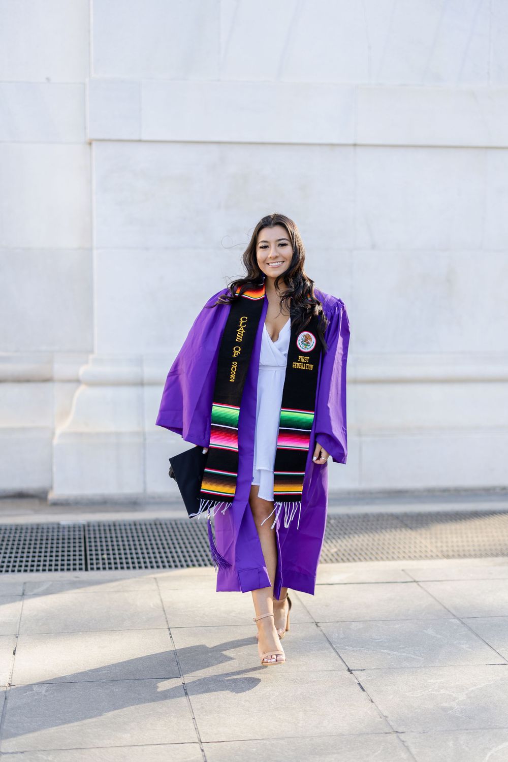 Graduate wearing purple gown and decorative stoles posing against white stone wall.