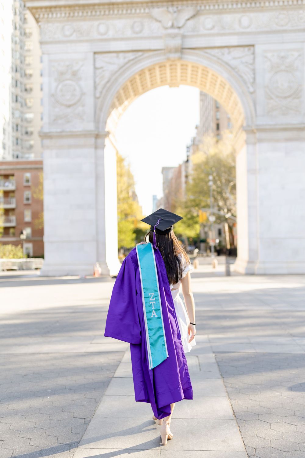 Graduate in purple cap and gown walks away from Washington Square Arch in NYC.