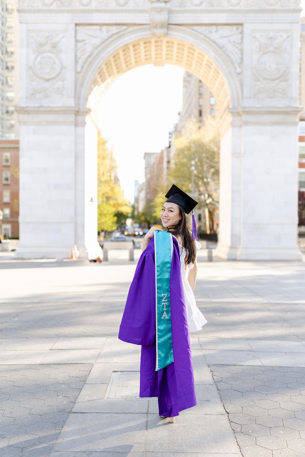 Graduate in purple regalia and turquoise stole posing under historic stone arch.