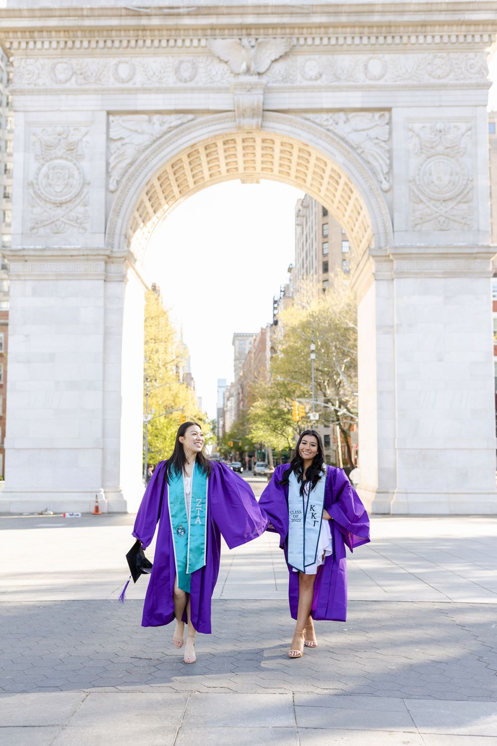 Two graduates walking together under Washington Square Arch in purple graduation robes.