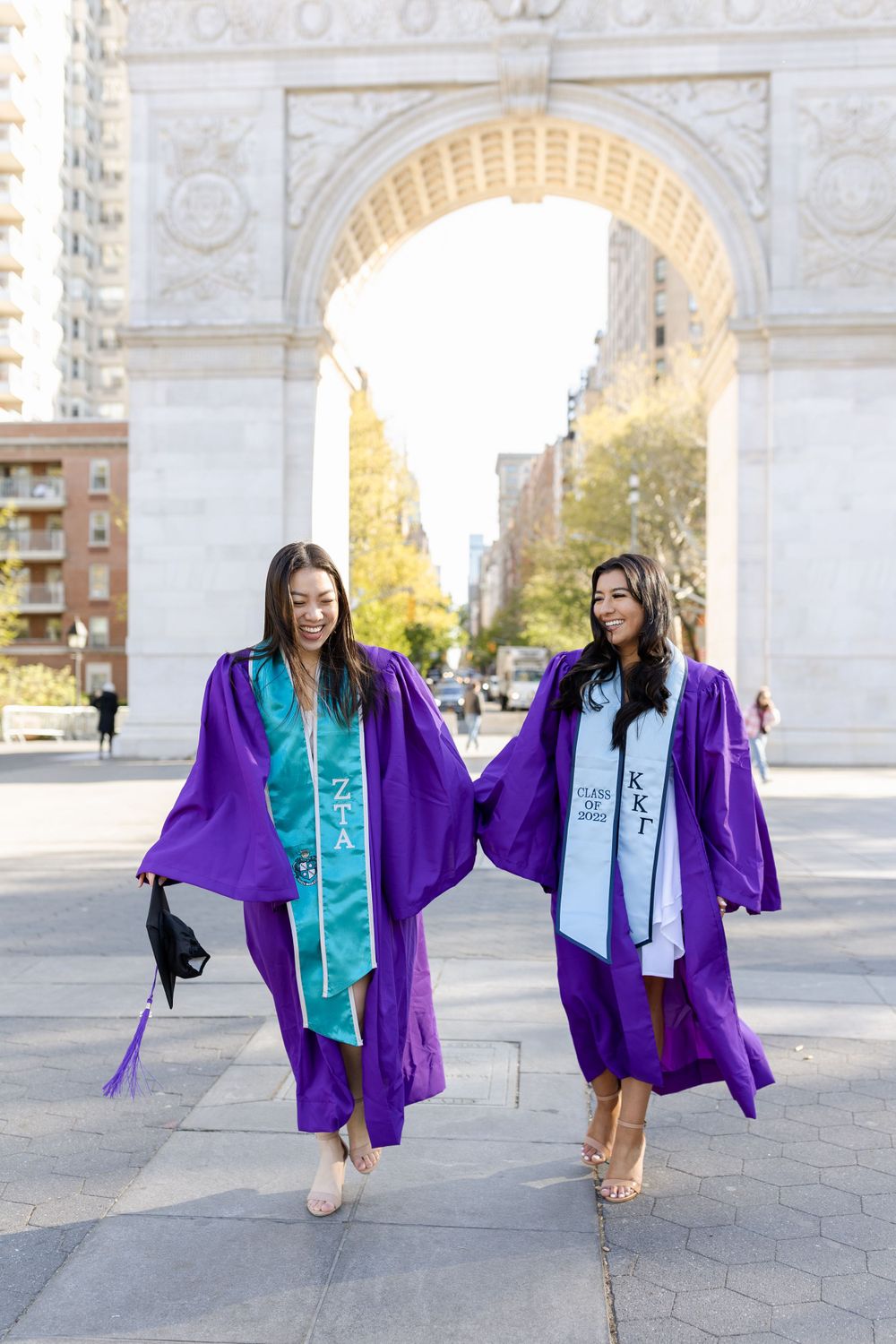 Graduates in purple robes walking and smiling near Washington Square Arch.