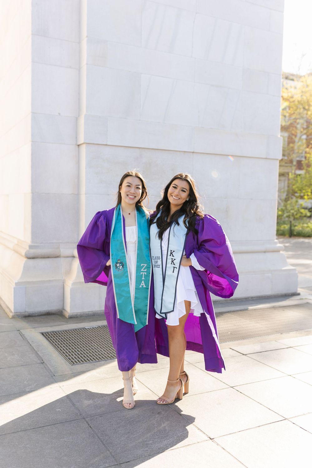 Graduates posing in purple gowns and teal stoles against white stone wall.