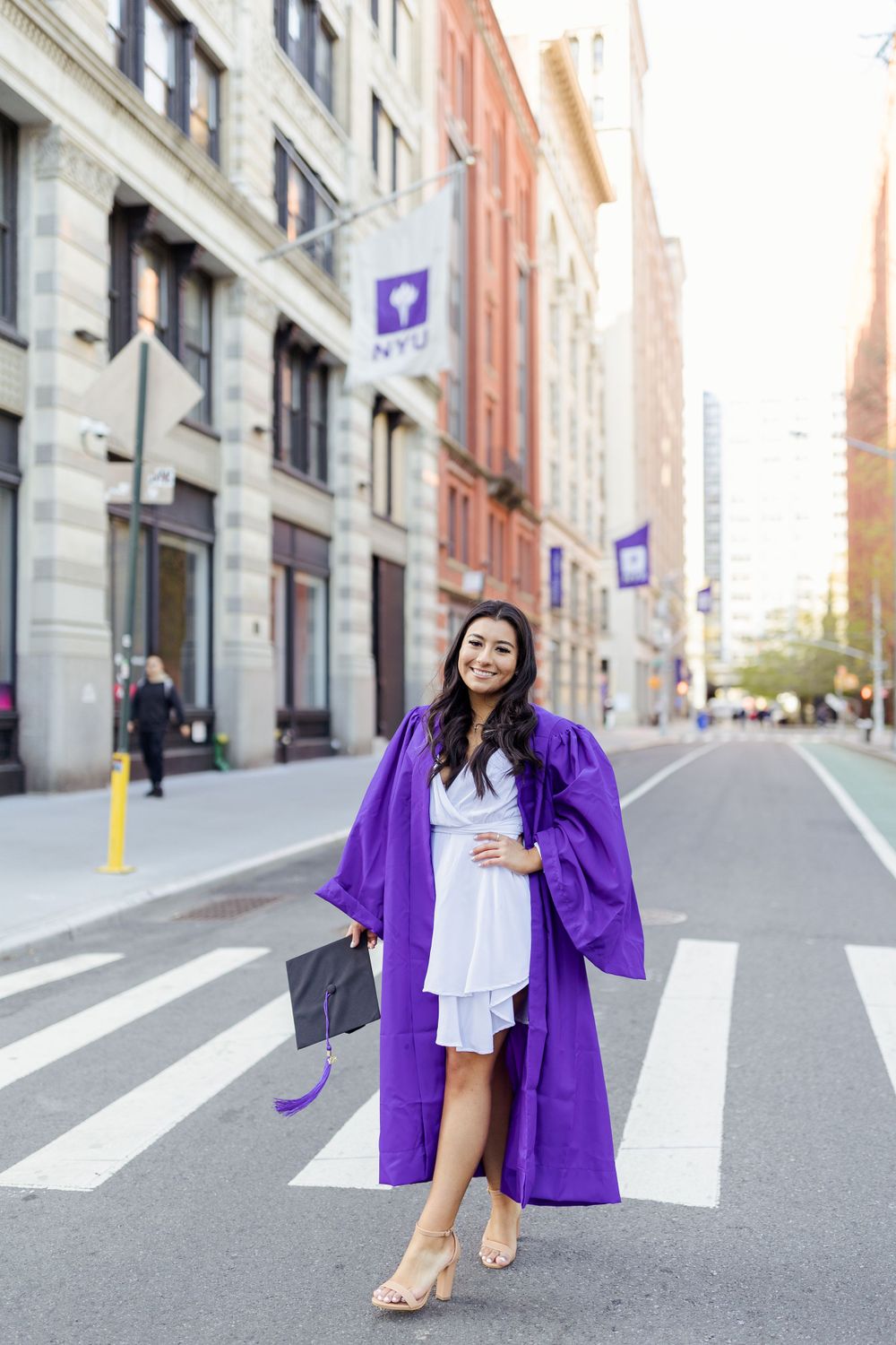 Graduate in purple gown poses on NYU campus street.
