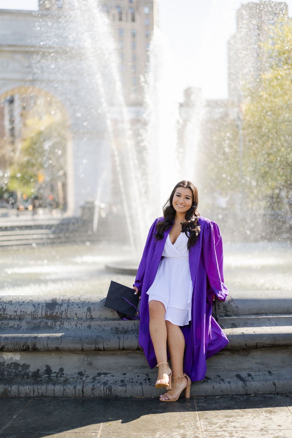 Graduate in purple gown poses by Washington Square Park fountain.
