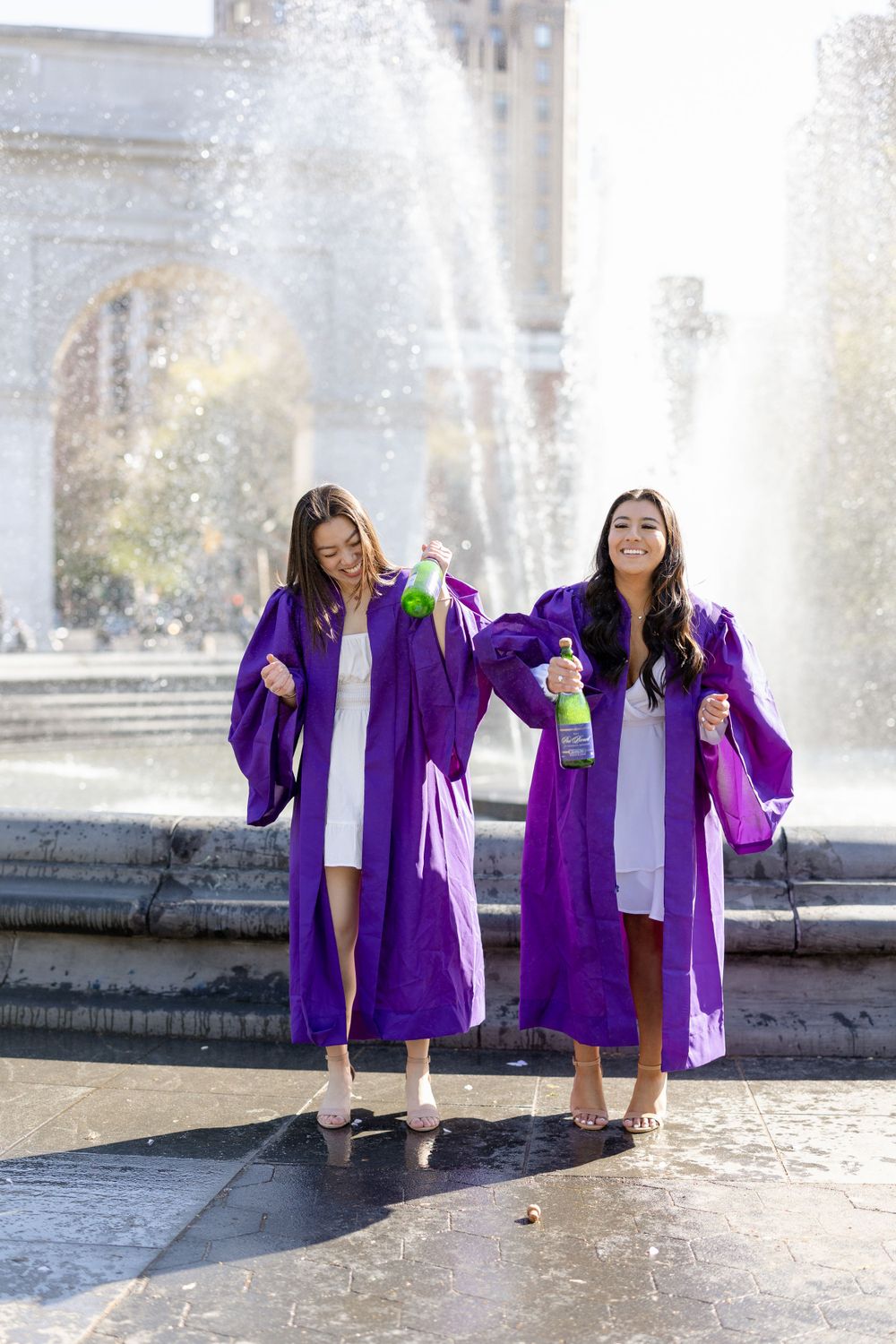 Two graduates celebrating with champagne by Washington Square Park fountain.