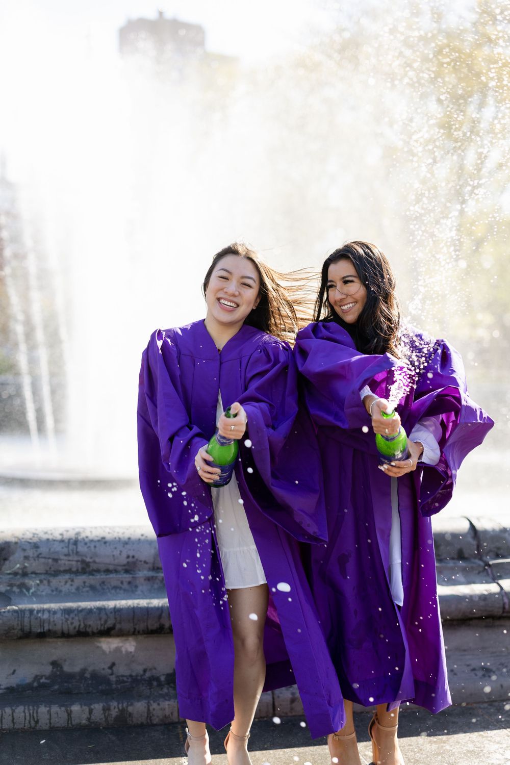 Two graduates in purple robes celebrate by fountains while holding bottles of champagne.
