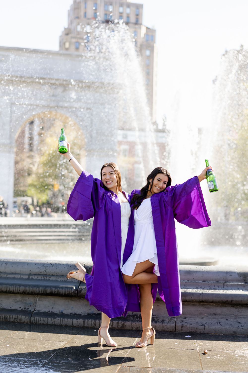 Purple-robed graduates pose with champagne bottles in front of Washington Square Arch fountain.
