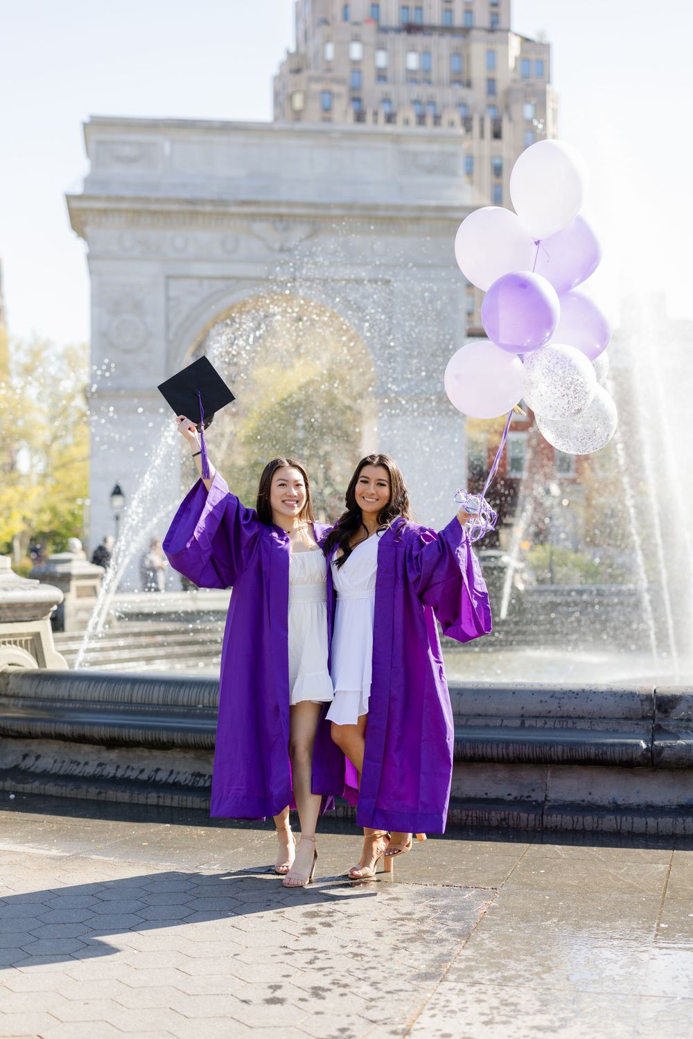 Graduates in purple robes hold balloons and caps by fountain during celebration photoshoot.