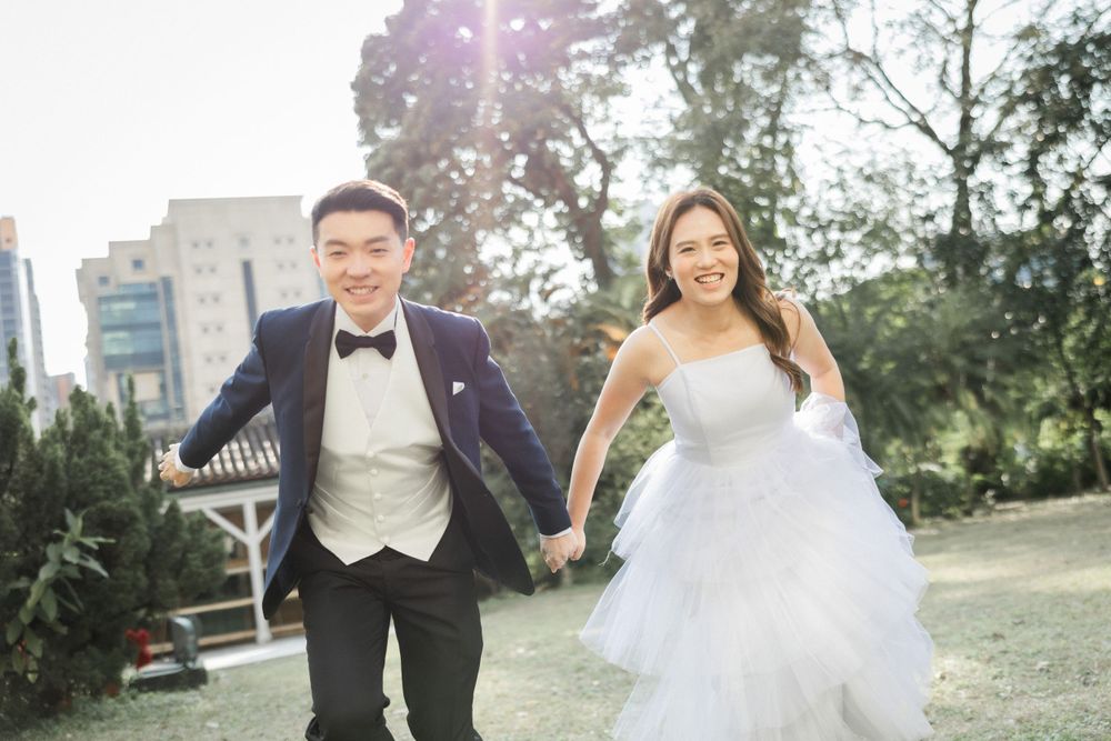 Smiling bride and groom run joyfully hand in hand through sunlit park with city buildings behind them.