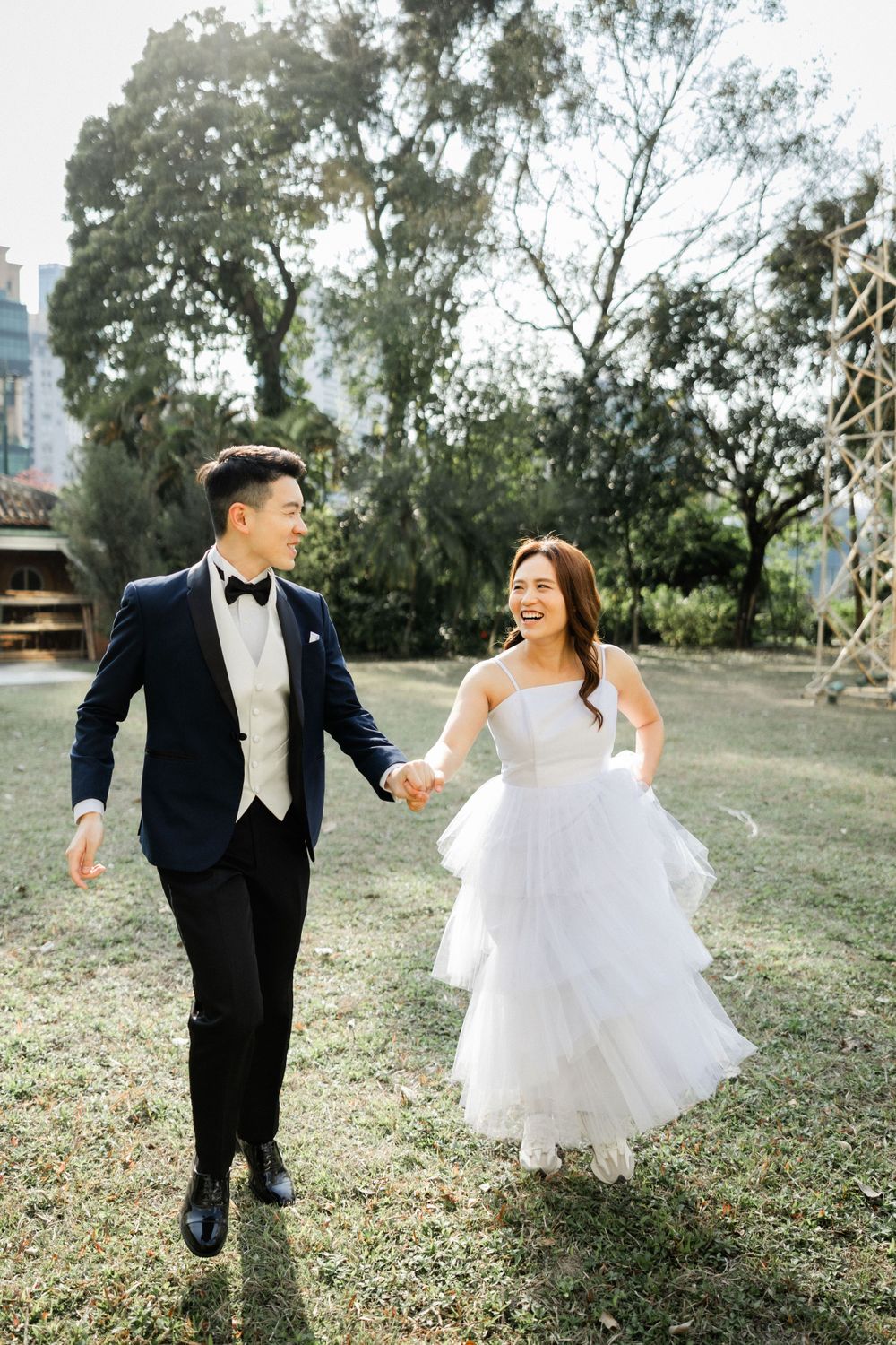 Happy couple laughs while walking hand in hand through green park with tall trees in soft sunlight.