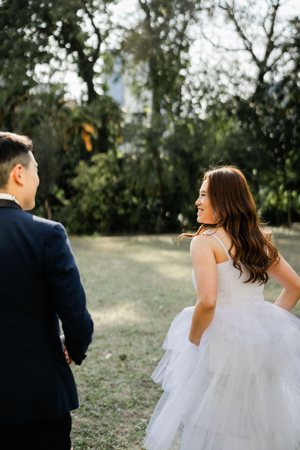 Smiling bride in layered white tulle gown glances back at groom while holding hands in garden setting.