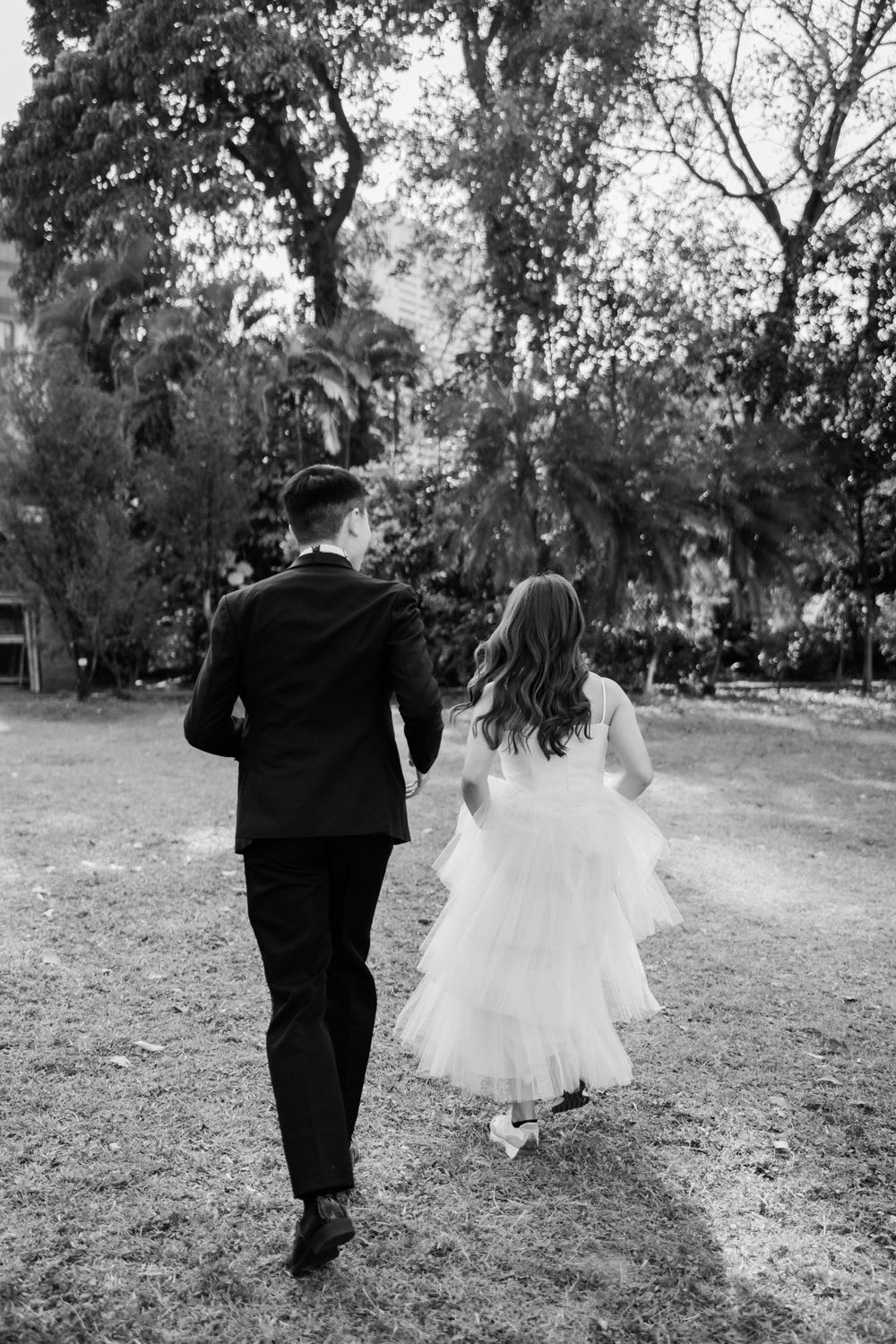 Black and white photo of bride and groom walking away hand in hand through a lush green park.