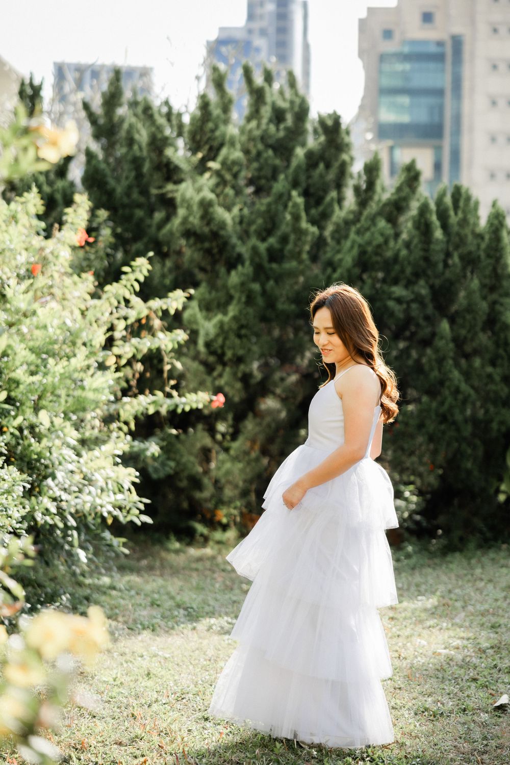 Elegant bride in layered white tulle gown stands gracefully beside tall evergreen trees in urban garden.