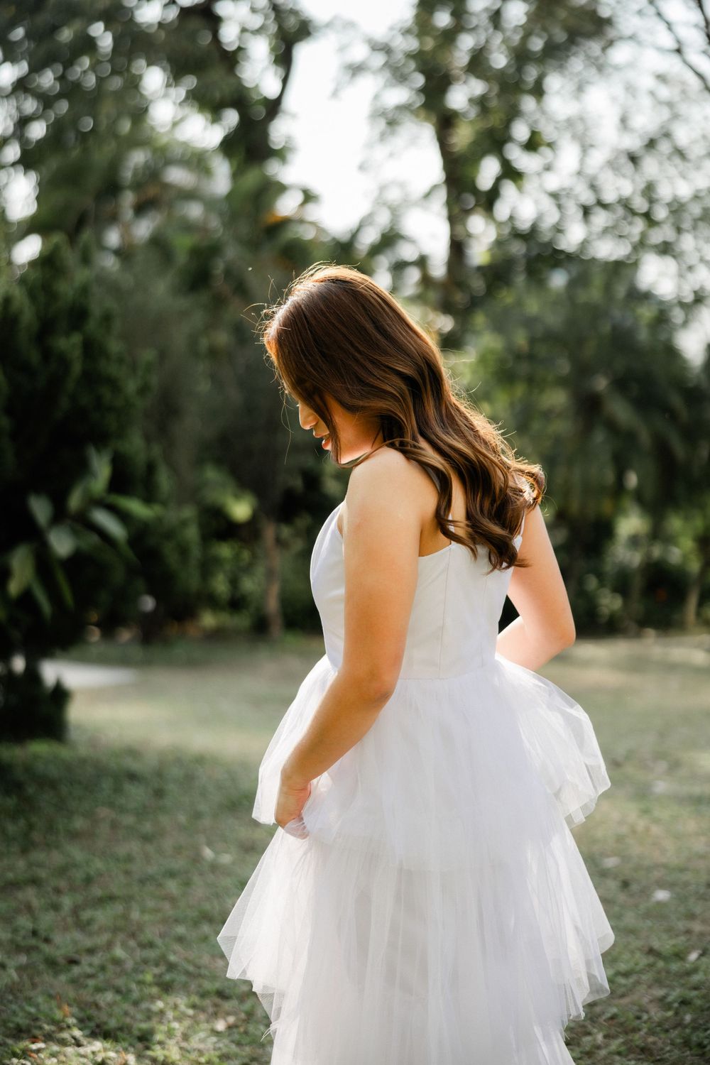 Bride in strapless layered white tulle gown looks down softly while standing in a lush green garden.