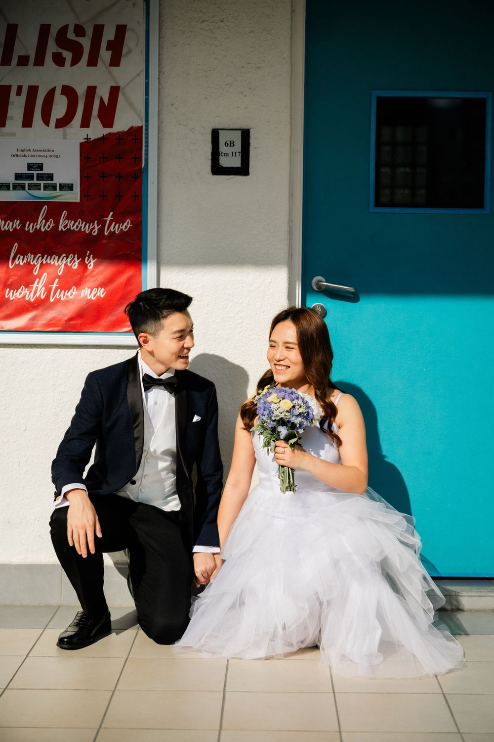 Bride and groom sitting together laughing outside a teal door, bride holding purple and white bouquet.