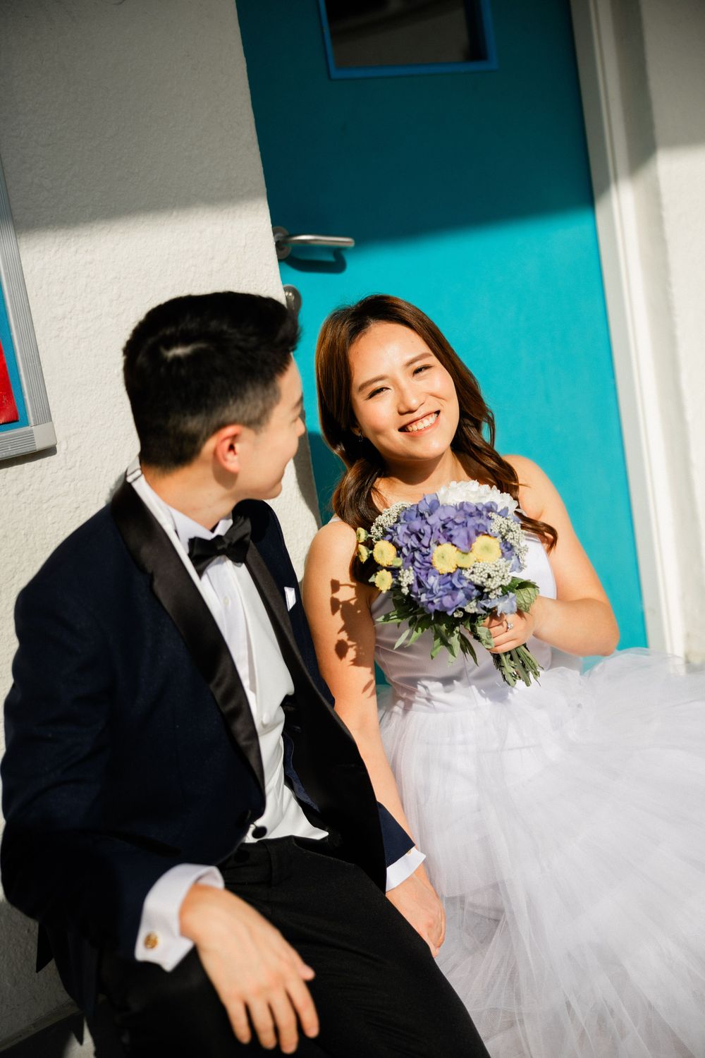 Smiling bride in white tulle gown holding purple bouquet gazes at groom in black tuxedo by teal door.