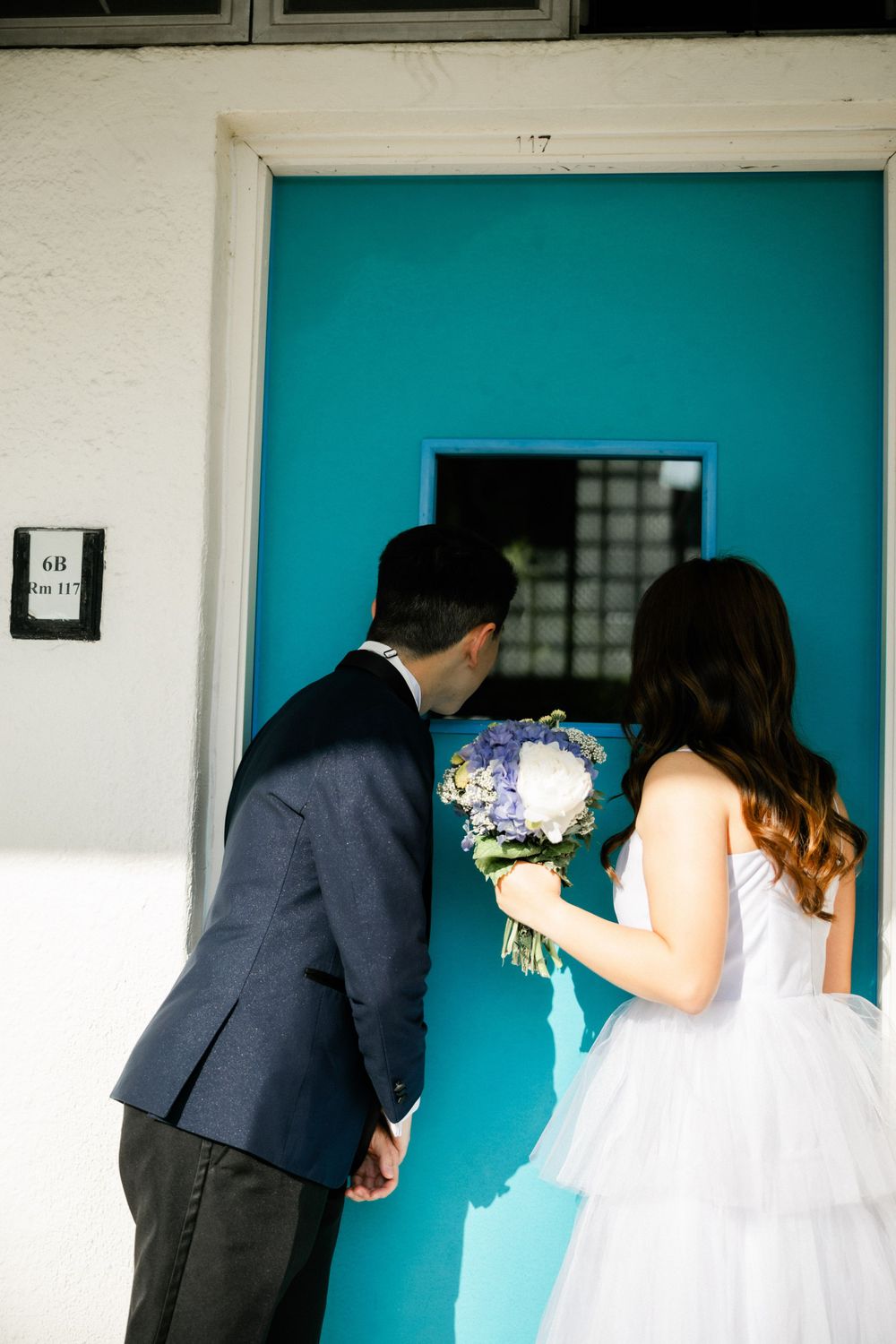 Bride and groom holding hands facing a vibrant teal door, bride carrying a purple and white bouquet.