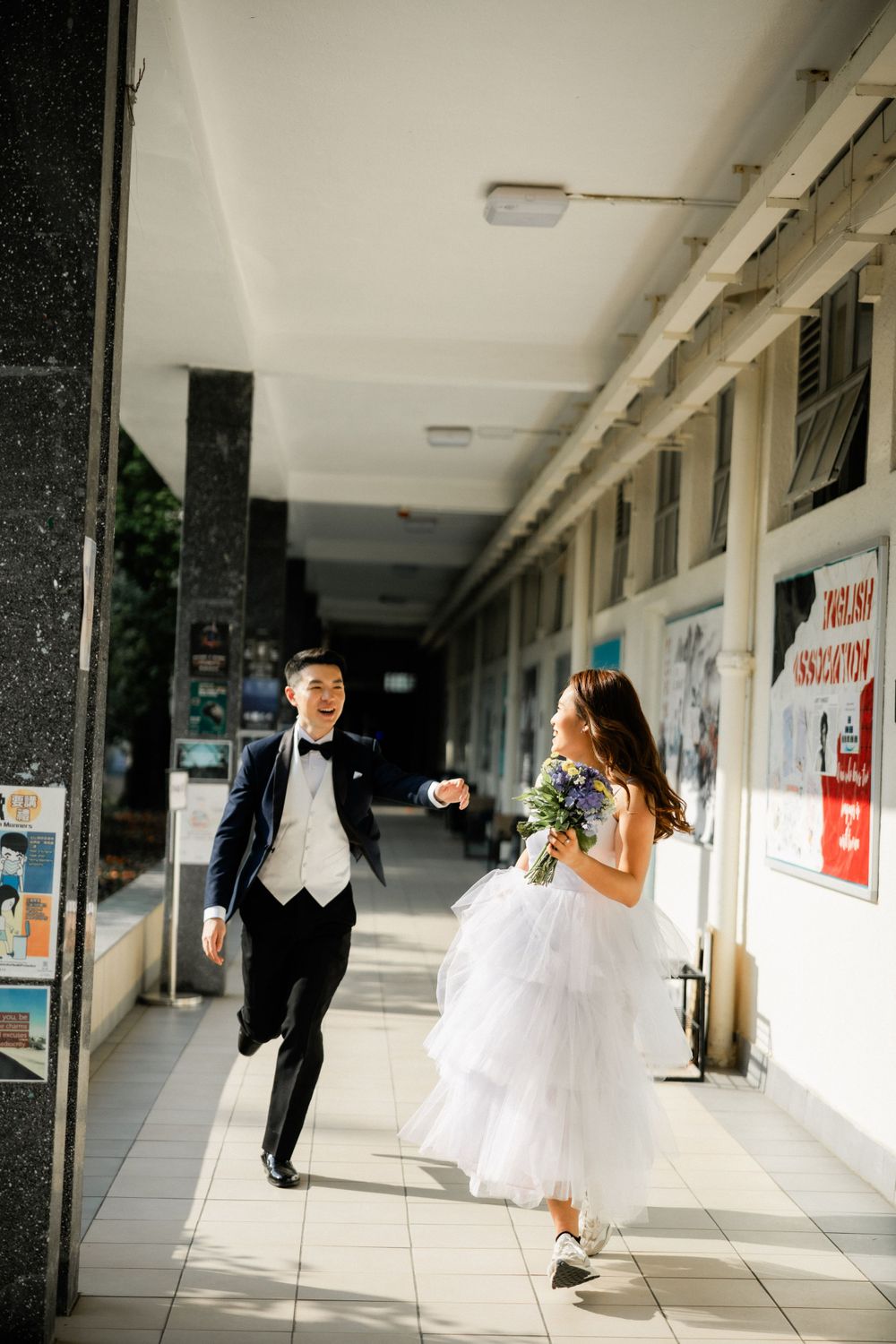 Bride and groom walking joyfully through a sunlit outdoor corridor, bride in layered white gown.