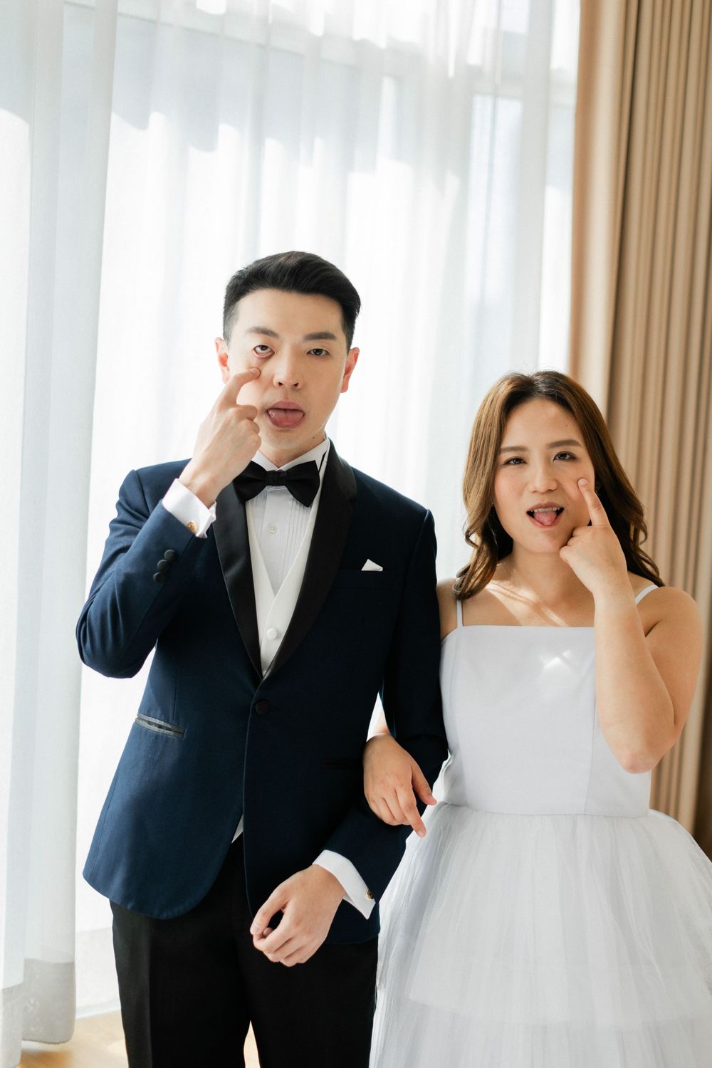Playful bride and groom in wedding attire making funny faces together by sheer curtains.