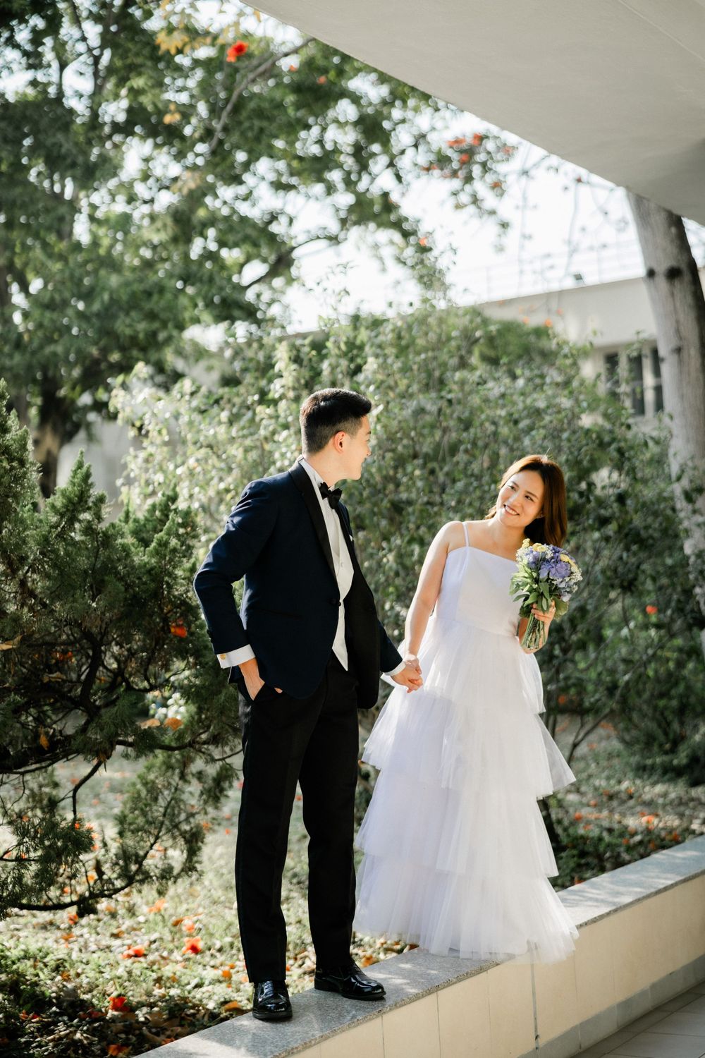 Bride and groom holding hands in a lush garden, bride smiling in white gown with purple bouquet.