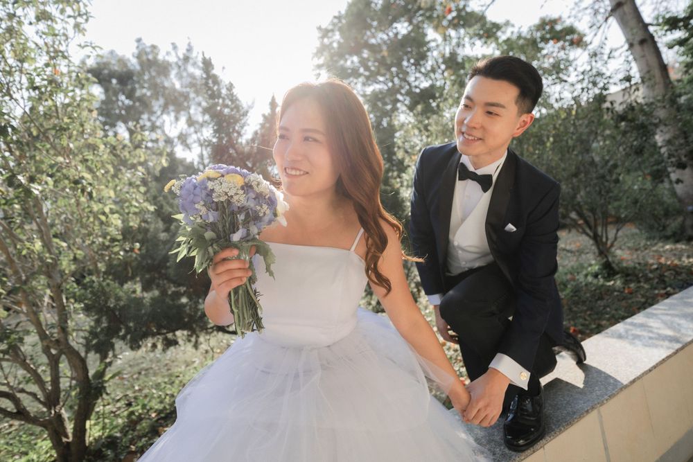 Bride and groom sitting on a ledge outdoors, bride holding purple bouquet with sunlight filtering through trees.