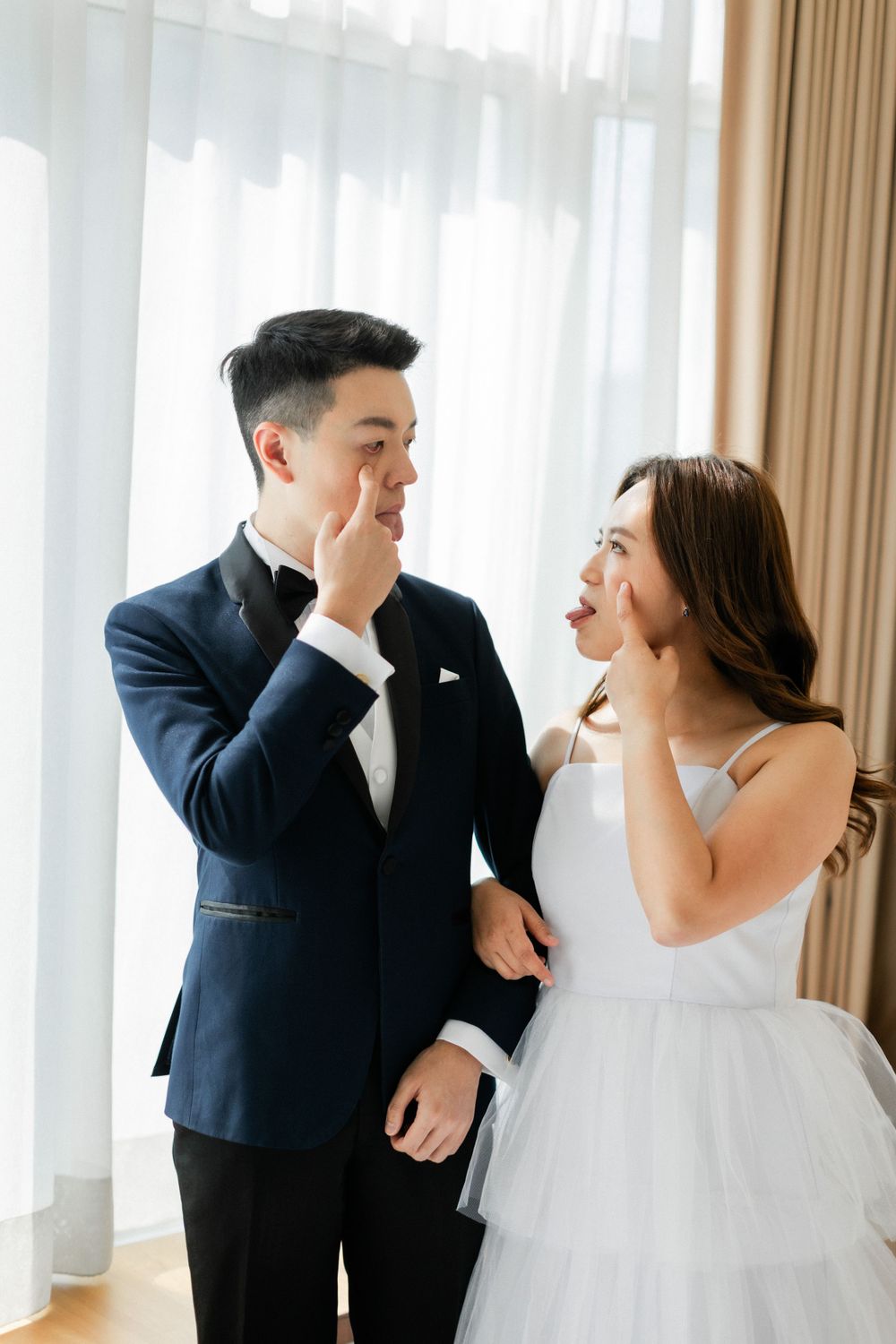 Bride and groom in wedding attire playfully touching each other noses while smiling indoors.