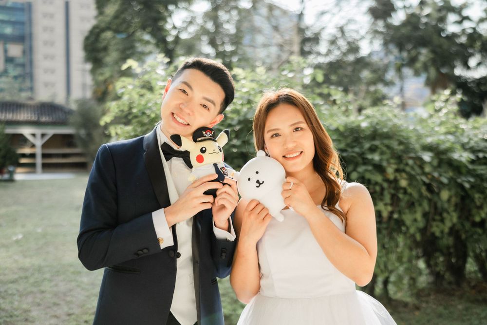 Smiling bride and groom holding cute stuffed animal toys in a sunny green park setting.