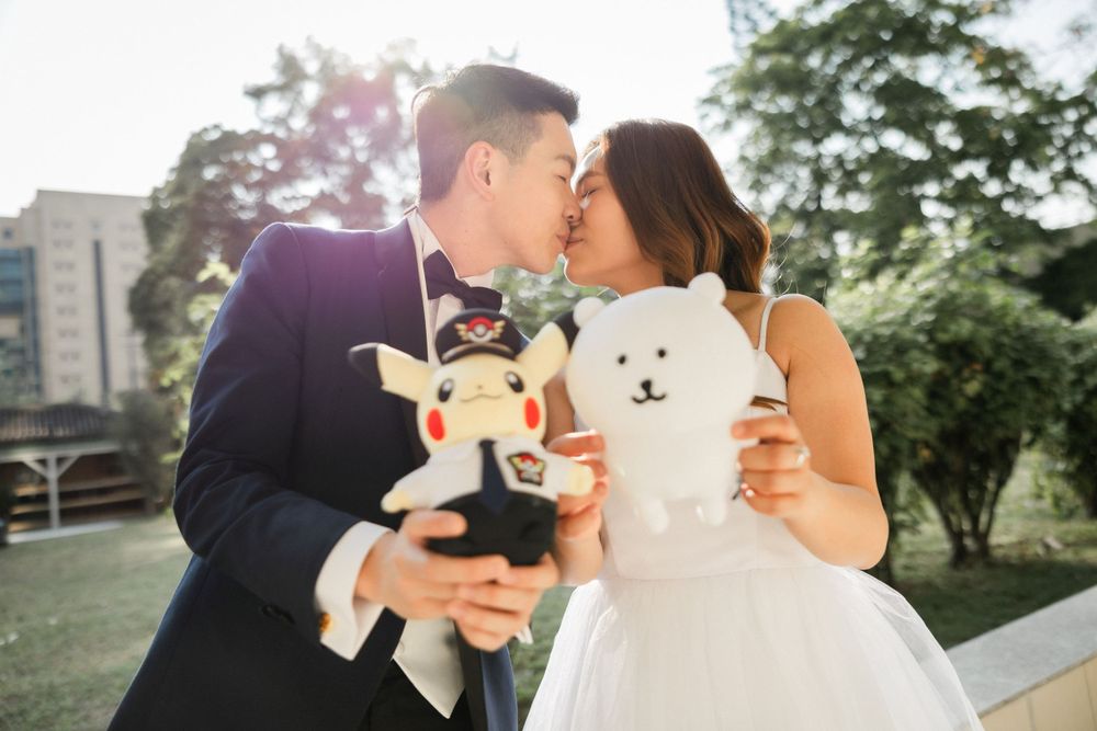 Bride and groom kissing while holding stuffed animal toys in a sunlit outdoor park.