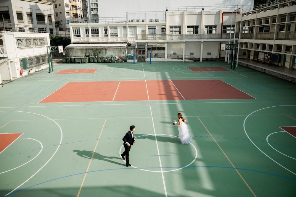 Bride in white gown and groom in tuxedo playing on a green and red rooftop basketball court.