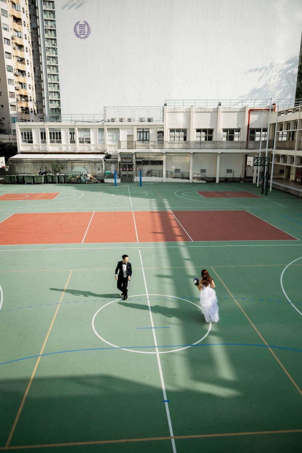 Bride in white gown and groom in black suit walking toward each other on a colorful outdoor basketball court.