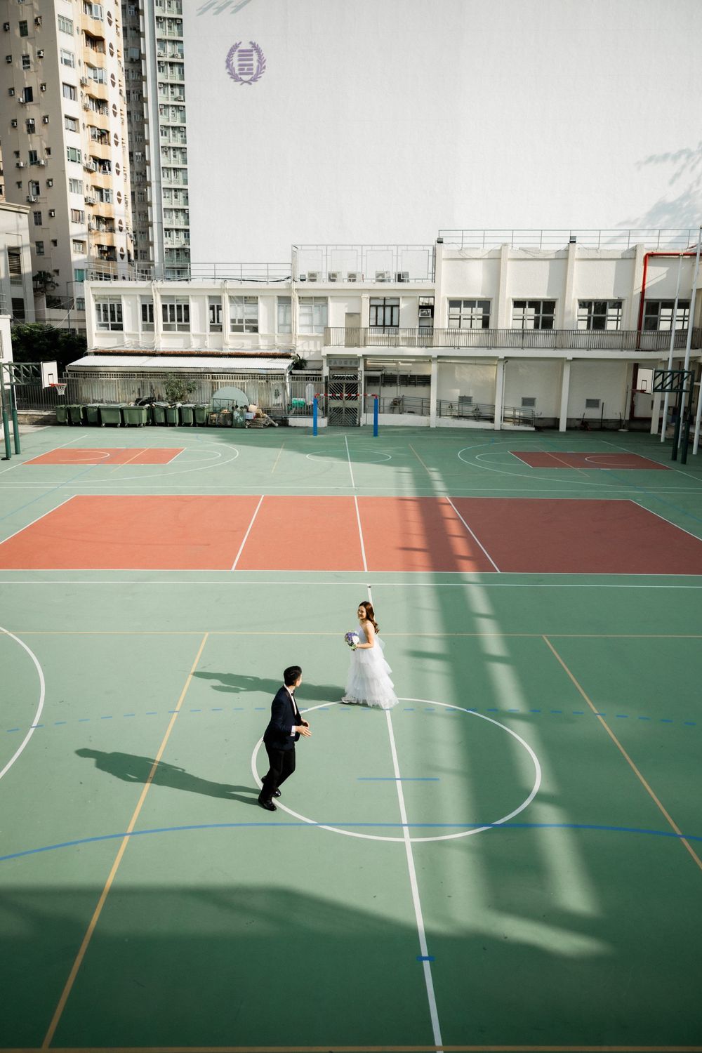 Wedding couple standing together on a green and red outdoor sports court with urban buildings in the background.