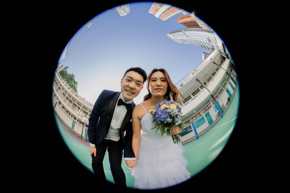 Asian bride and groom posing together in a fisheye lens photo, bride holding a purple and white bouquet.